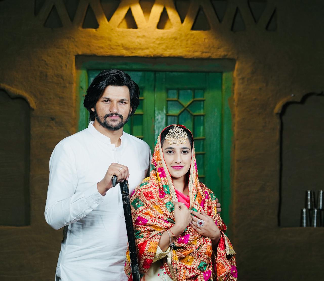 A couple stands elegantly in colorful traditional attire, showcasing cultural heritage indoors.