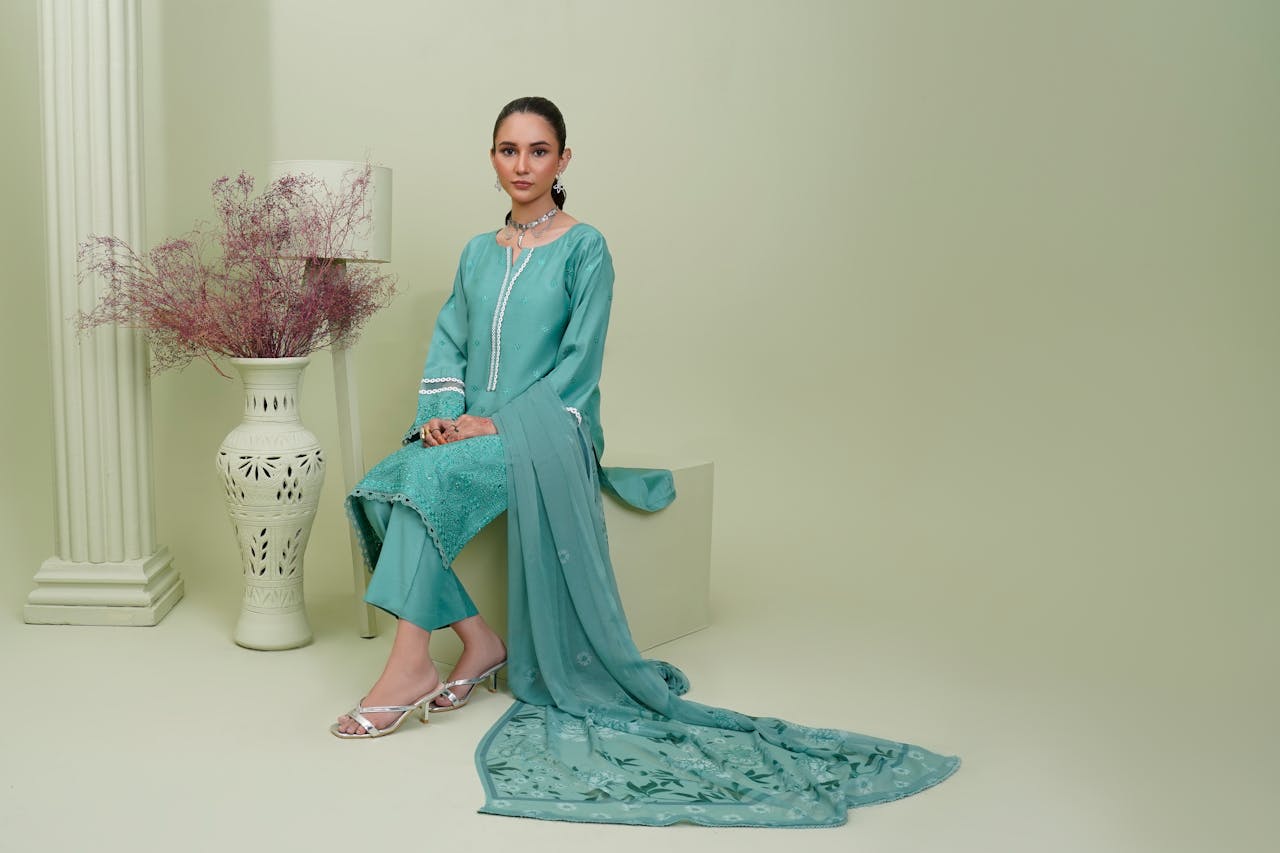 South Asian woman in an embroidered teal dress posing indoors, showcasing fashion and elegance.